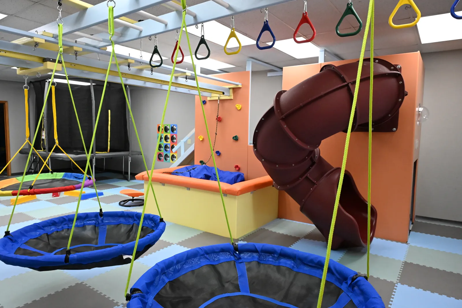 Platform swings hanging from a ceiling ladder with colorful rings, beside a tube slide and climbing wall in the sensory gym