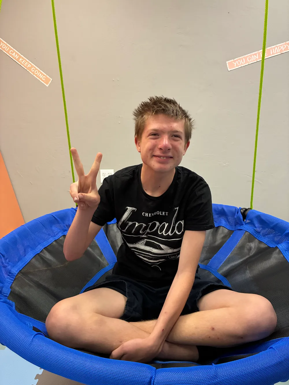 A smiling teen sitting cross-legged on a blue platform swing, flashing a peace sign
